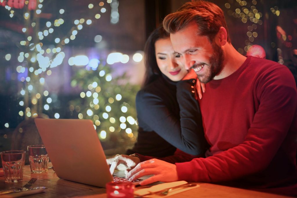 Ein Paar, das in einem gemütlichen Restaurant mit festlichen Bokeh-Lichtern einen Laptop benutzt und gemeinsam online einkauft.